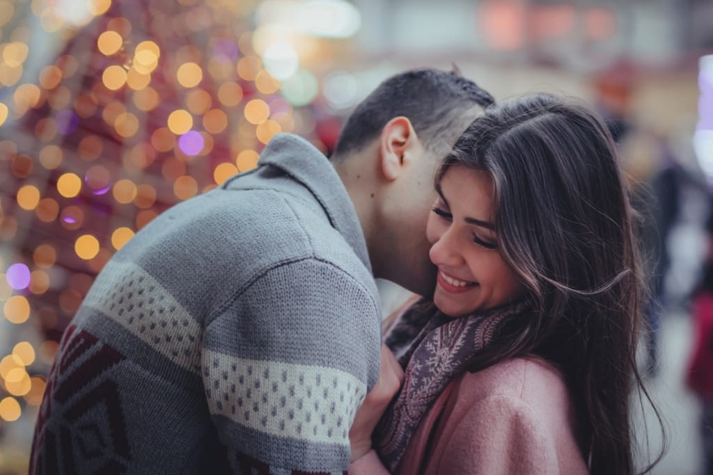 man kisses a woman on the cheek with Christmas tree in the background in winter